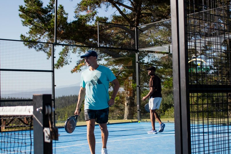 Açık havadaki bir padel kortunda ellerinde raketlerle yürüyen iki oyuncu, camın arkasında ağaçlar